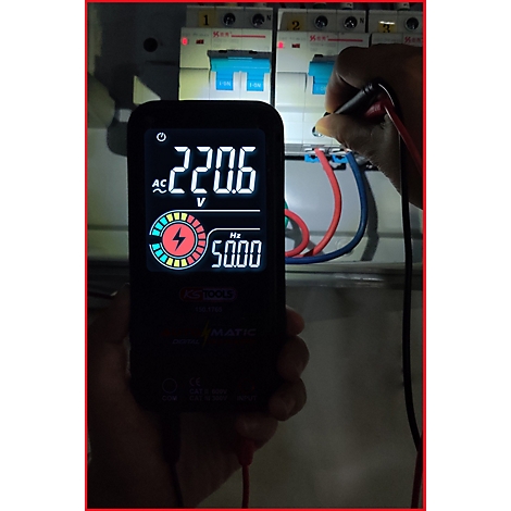 Multimeter zeigt 220,6 V und 50,00 Hz. Eine Hand berührt einen Stromkreis. Im Hintergrund sind Sicherungsautomaten zu sehen.